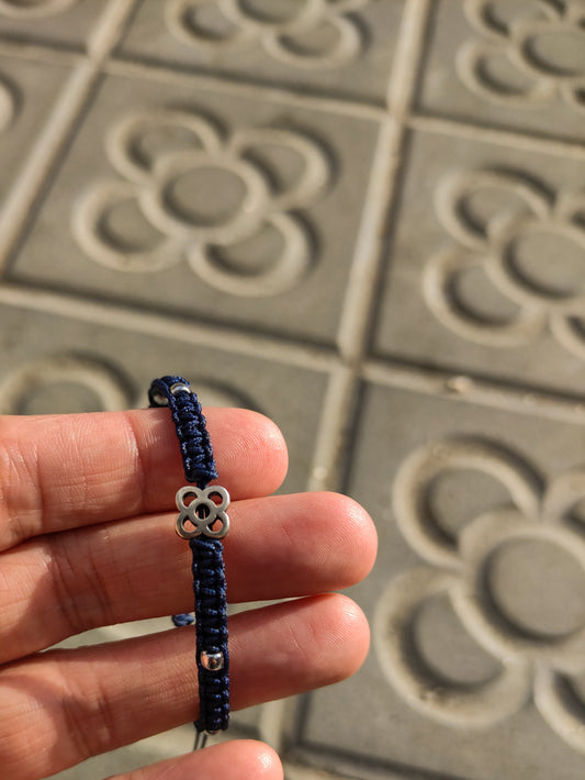 Pulsera de macramé con símbolo del panot en una mano que lo enseña y se ven baldosas de la ciudad de Barcelona con el mismo símbolo